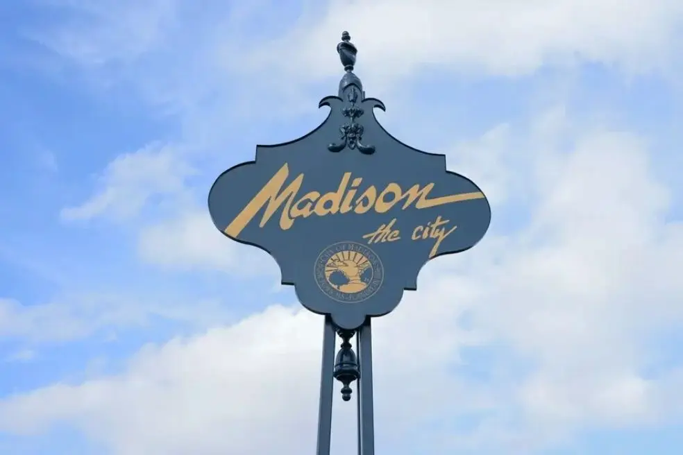 A decorative black sign against a blue sky reading Madison the city above the City of Madison, Mississippi municipal seal.