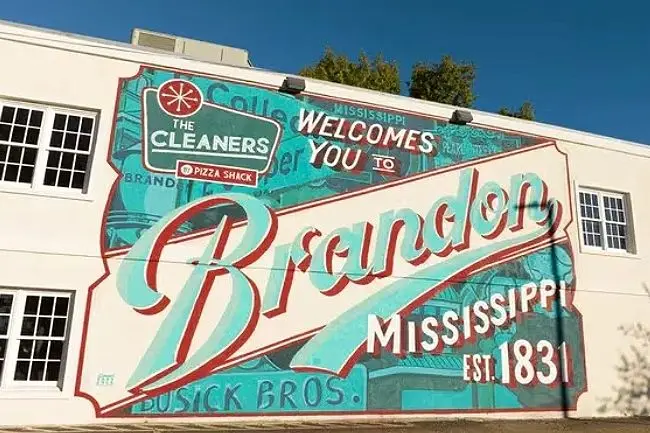 A large mural on the side of a building that reads Mississippi Welcomes You To Brandon, Mississippi Est. 1831, featuring logos for The Cleaners and Pizza Shack.