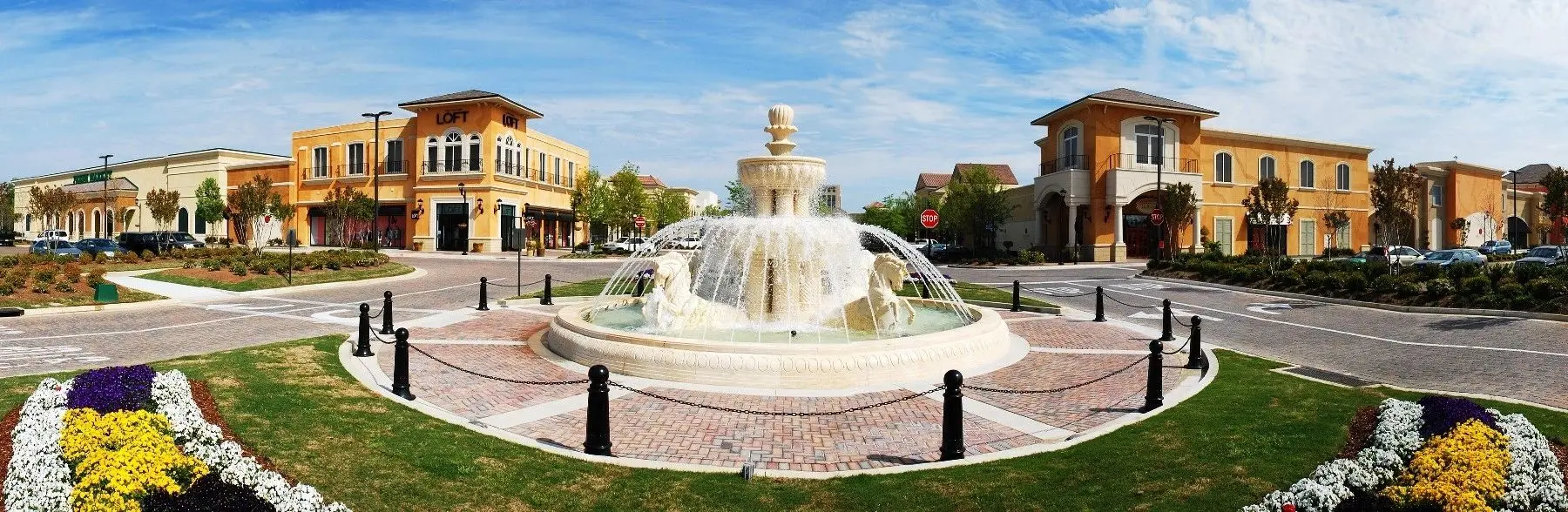 A wide-angle view of a tiered stone fountain in the center of an outdoor shopping plaza surrounded by storefronts.