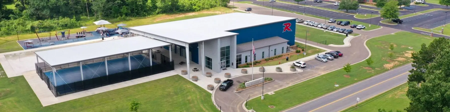 Aerial view of a community recreation center with a blue gym building, outdoor sports courts, a playground, and an adjacent parking lot.