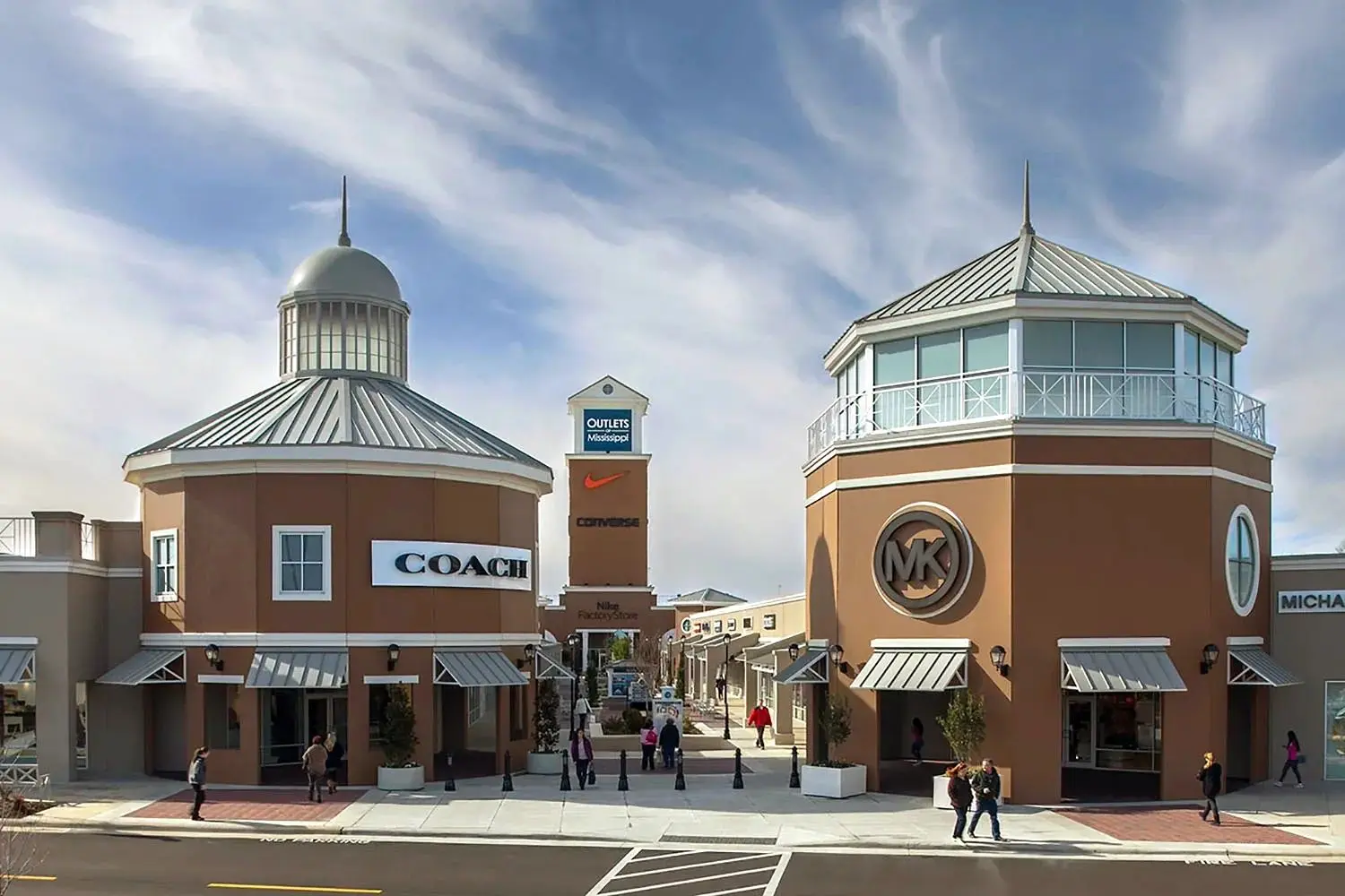 An outdoor shopping center with Coach and Michael Kors storefronts flanking a central walkway, featuring a tall clock tower in the background labeled Outlets Mississippi.
